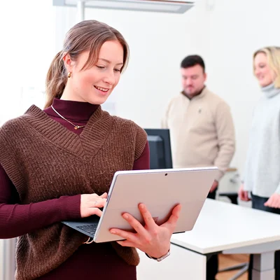 Eine WOBAU-Mitarbeiterin steht mit ihrem Tablet im Büro, sie tippt. Im Hintergrund sind zwei Kollegen zu sehen, wie sie an einem Schreibtisch stehen und auf einen Computer schauen.