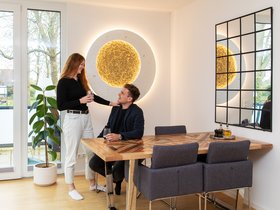 Ein Mieterpaar im Esszimmer ihrer Wohnung. Der Mann sitzt, die Frau steht neben ihm und legt eine Hand auf seine Schulter. Beide schauen und lächeln sich an. Beide haben ein Glas Wasser in der Hand.