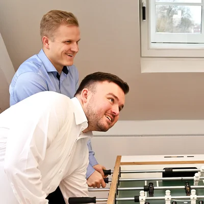 Vier Mitarbeiter (drei Männer, eine Frau) stehen im Aufenthaltsraum am Tischkicker und spielen. Alle sind gut gelaunt und lächeln sich an.