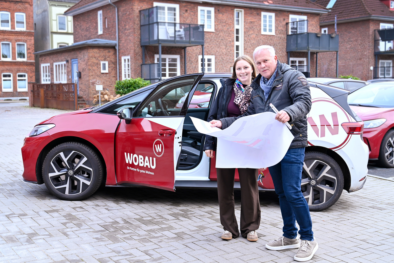 Zwei WOBAU-Mitarbeitende (ein Mann und eine Frau) stehen vor einem WOBAU-Auto. Der Mann hält einen Bebauungsplan vor sich, die Frau steht daneben. Beide Personen schauen lächelnd in die Kamera.