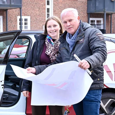 Zwei WOBAU-Mitarbeitende (ein Mann und eine Frau) stehen vor einem WOBAU-Auto. Der Mann hält einen Bebauungsplan vor sich, die Frau steht daneben. Beide Personen schauen lächelnd in die Kamera.